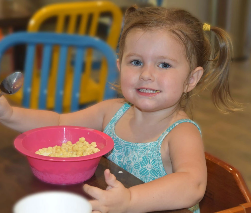 young girl smiling and holding spoon as she eats a bowl of cereal